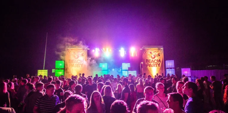 Viele Gäste stehen bei Nacht vor einer Bühne mit zwei Beachparty-Bannern und leuchtenden quadratischen Elementen in bunten Farben.