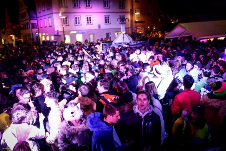 Eine große kostümierte Menschenmenge füllt nachts einen Platz vor einem beleuchteten Gebäude; violettes Licht fällt auf Besucher und Zelte.
