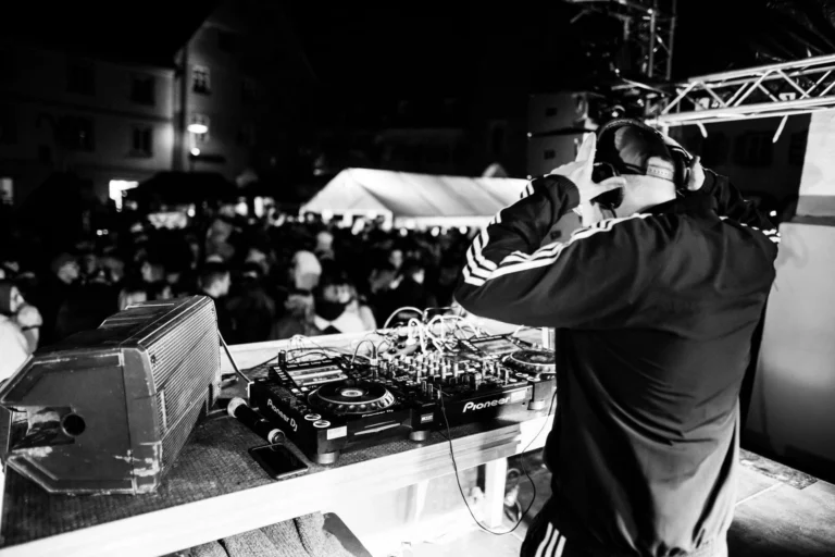 Ein DJ mit Kopfhörern steht am Mischpult einer Außenbühne und schaut auf ein dichtes Publikum vor den Gebäuden.