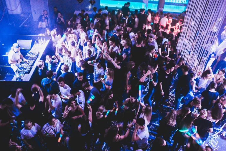 Eine dicht stehende Menschenmenge füllt den Club neben einem DJ-Bereich; Besucher heben die Arme unter blauen und violetten Lichtern.