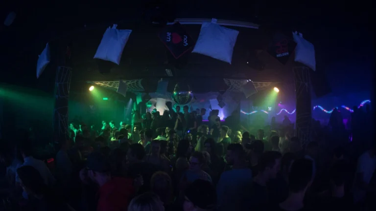 Eine volle Tanzfläche liegt im dunklen Club unter blauen und grünen Scheinwerfern und von der Decke hängenden weißen Flächen.