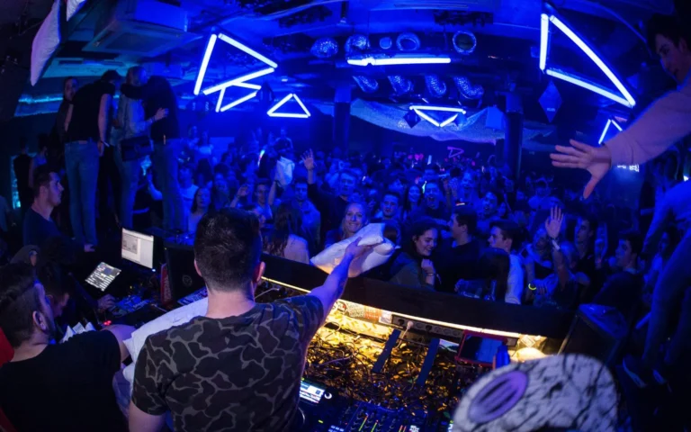 Ein DJ steht mit dem Rücken zur Kamera am Pult und breitet die Arme aus; vor ihm tanzt eine Menge unter blauen dreieckigen Leuchten.