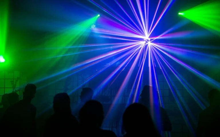 Blaue Laserstrahlen fächern sich sternförmig über dunklen Gästen auf einer grünen und blauen Tanzfläche auf.