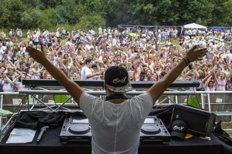 Ein DJ im weißen T-Shirt und schwarzer Kappe breitet hinter dem Pult beide Arme vor einer großen Festivalmenge aus.