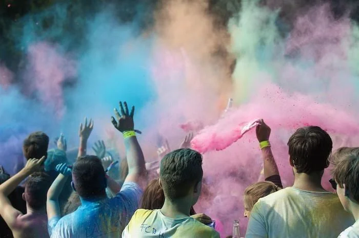 Festivalgäste heben vor blauen, gelben und pinken Pulverwolken die Hände; mehrere haben Farbe auf Haaren und Kleidung.