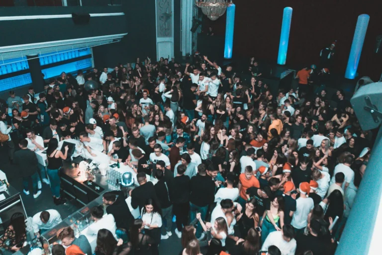 Eine dicht stehende Menschenmenge füllt einen Club mit Bar und hohen hellen Säulen; blaue Lichter beleuchten die Wände.