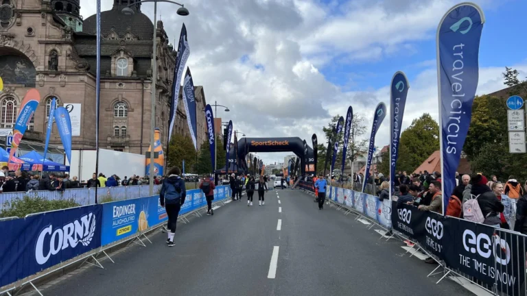 Eine breite Straße ist mit Gittern und blauen Werbebannern eingerahmt vor einem schwarzen Bogen und zahlreichen Fahnen.