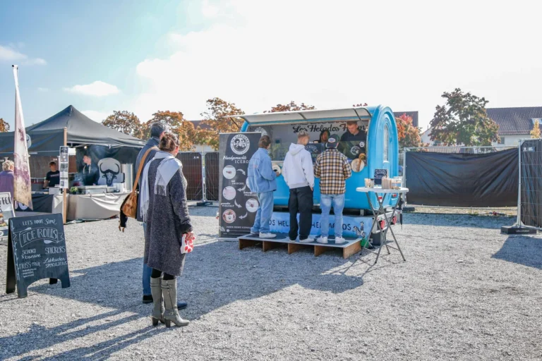 Eine Frau steht auf dem Schotterweg vor einem blauen Eiswagen und einem schwarzen Verkaufszelt beim Streetfood-Festival Puchheim.