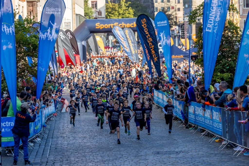 Viele Läufer warten auf der abgesperrten Straße unter einem schwarzen SportScheck-Startbogen; Zuschauer stehen am Rand.