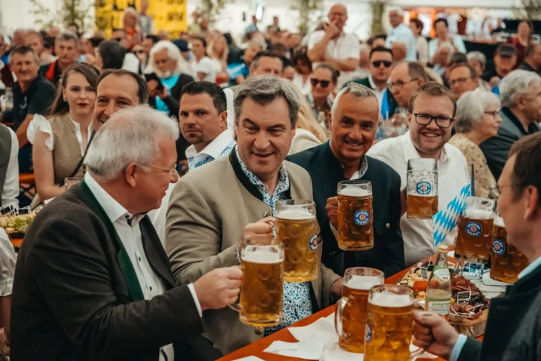 Markus Söder sitzt mit mehreren Gästen am Biertisch und hält einen großen Bierkrug; ringsum stehen und sitzen weitere Besucher.