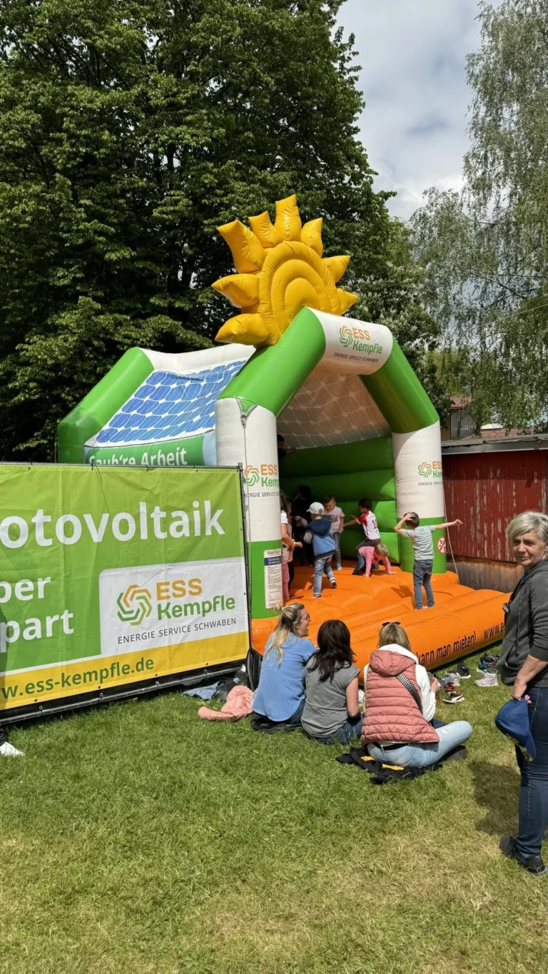Eine grün-orange Hüpfburg mit großer gelber Sonnenfigur steht auf der Wiese neben einem grünen Werbeschild und Besuchern.