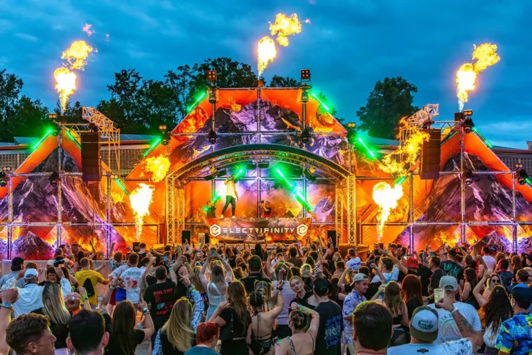 Große Flammen steigen über der bunt beleuchteten Festivalbühne auf; davor steht eine dichte Menge unter dunklem blauem Himmel.