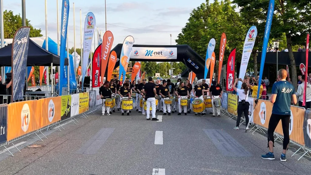 Eine Gruppe steht auf der abgesperrten Straße vor einem schwarzen M-Net-Startbogen; bunte Fahnen und orange Banner säumen den Bereich.