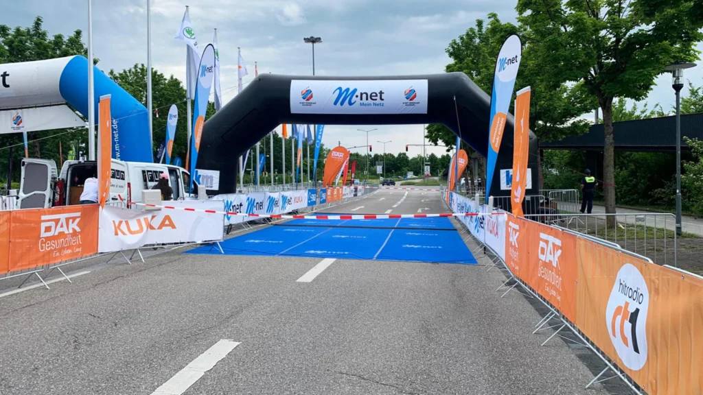 Ein schwarzer M-Net-Startbogen steht über einer Straße mit blauen Bodenmatten; orangefarbene Banner und Fahnen säumen die Strecke.