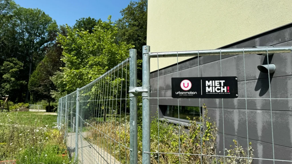 Ein Metallbauzaun mit schwarzem Urbanmotion-Mietschild steht vor der grauen unteren Fassade eines hellgrünen Gebäudes.