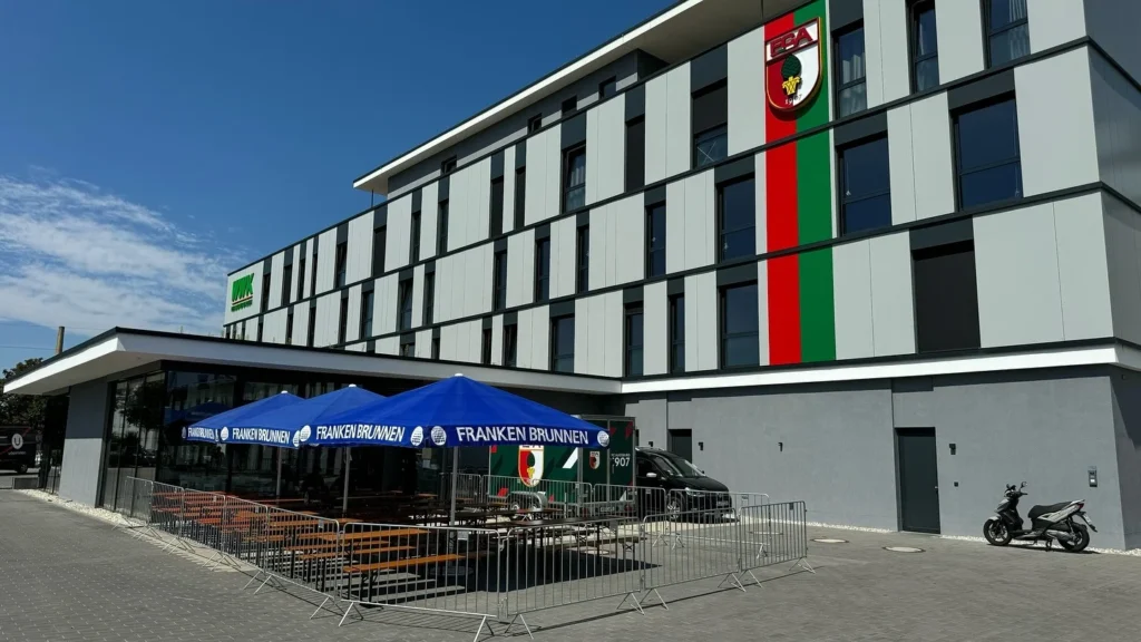 Ein mehrstöckiges Gebäude mit weiß-schwarzer Fassade und grün-rotem Streifen steht neben einem abgesperrten Terrassenbereich.