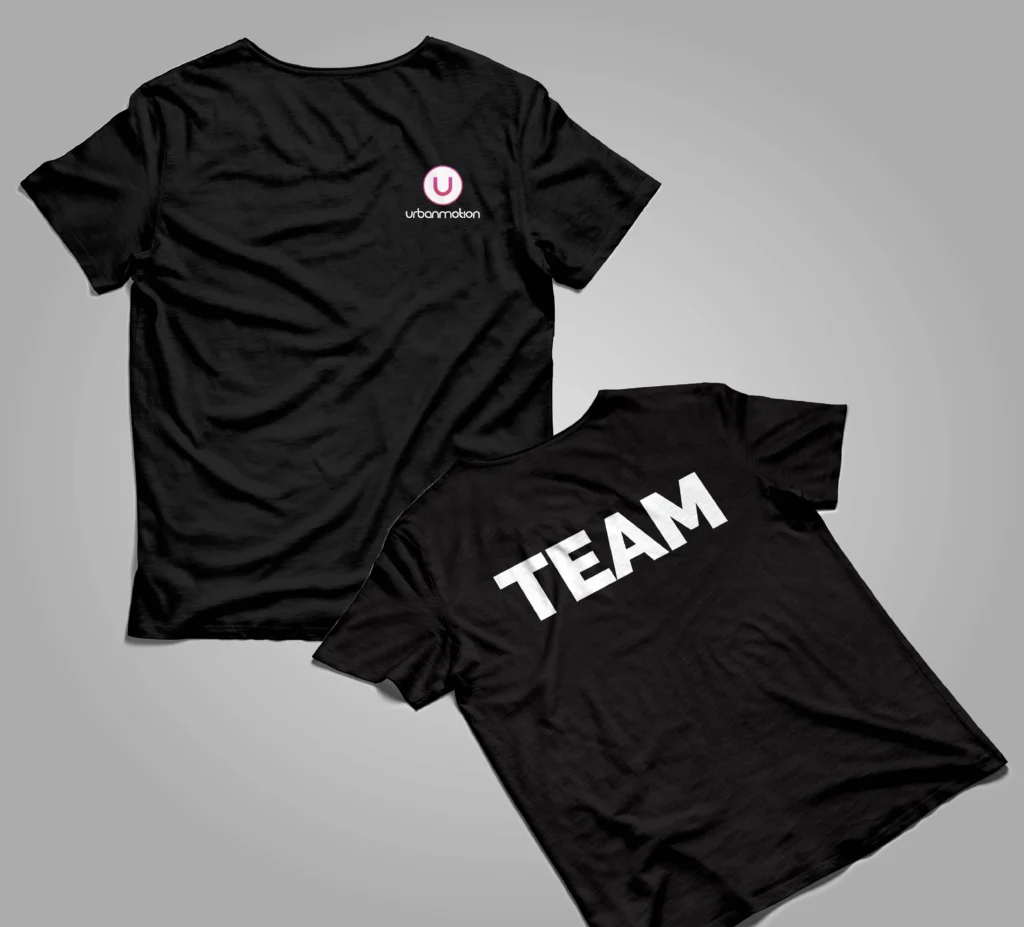 Zwei schwarze T-Shirts liegen auf grauem Hintergrund; eines zeigt ein kleines Urbanmotion-Logo, das andere den weißen Schriftzug Team.