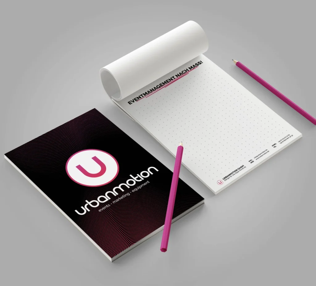 Ein schwarzer Notizblock mit Urbanmotion-Logo liegt neben einem geöffneten weißen Block; zwei pinke Stifte liegen darüber.
