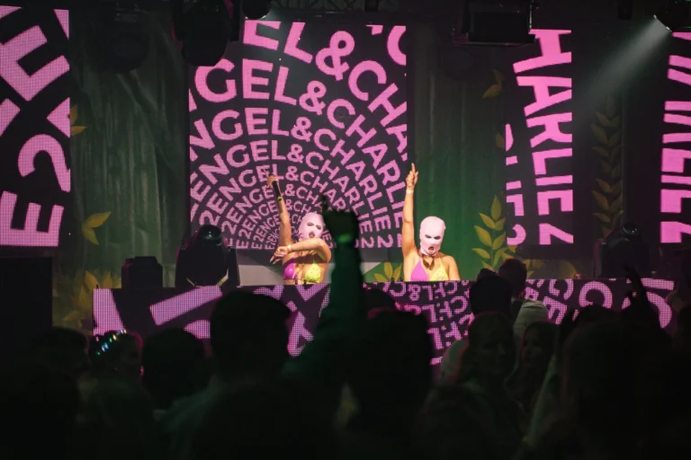 Zwei DJs mit pinken Masken stehen am Pult vor großen pinken Schriftzügen; dunkle Silhouetten des Publikums füllen den Vordergrund.