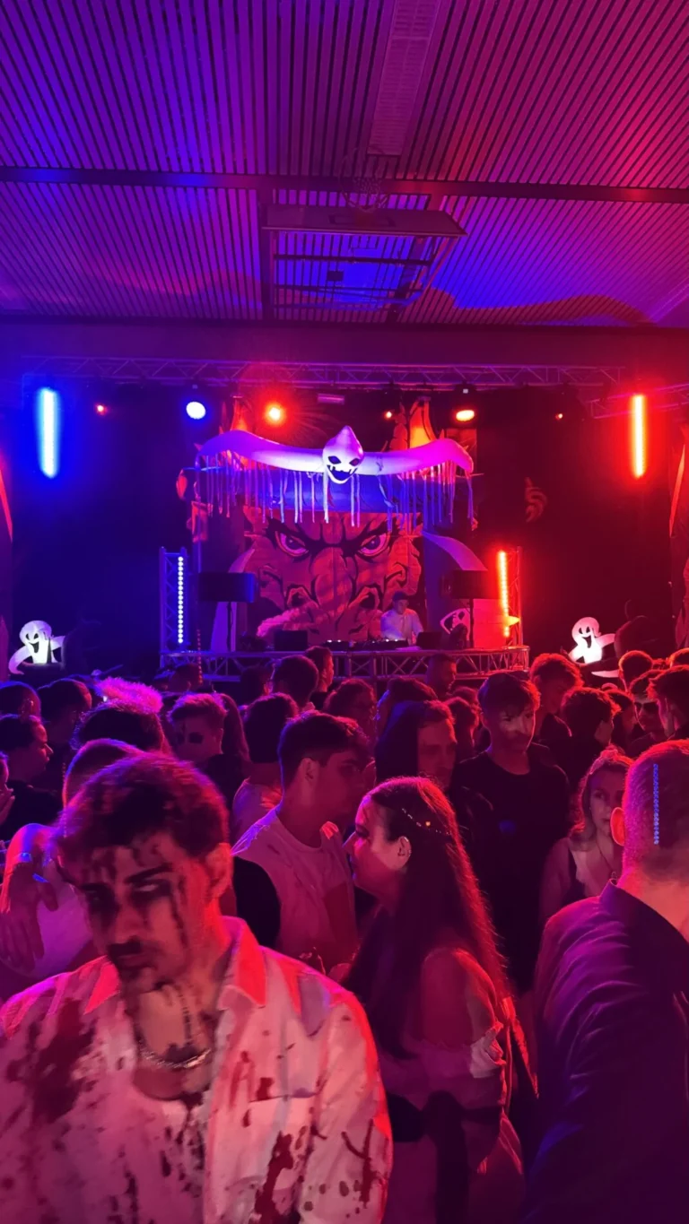 Viele Gäste stehen dicht vor einer blau und pink beleuchteten Bühne; vorn trägt ein Besucher ein helles gemustertes Hemd.