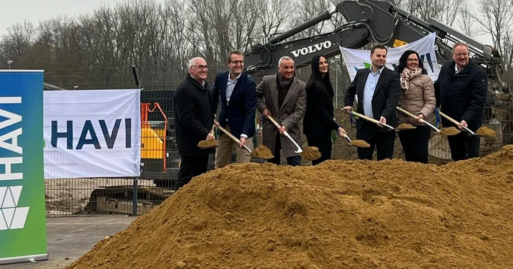 Eine Gruppe hält Spaten hinter einem Erdhügel; ein Bagger und HAVI-Banner stehen im Hintergrund.