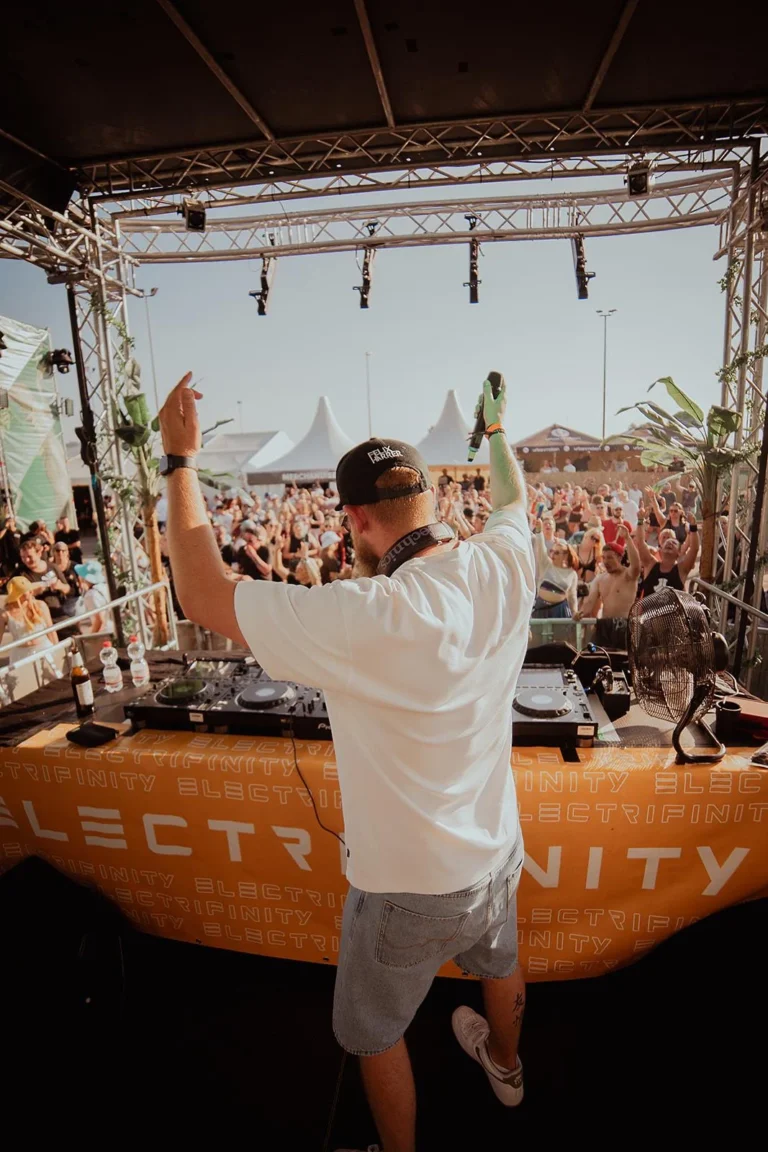 Ein DJ im weißen Shirt steht mit dem Rücken zur Kamera und hebt beide Arme vor einem vollen Festivalpublikum.