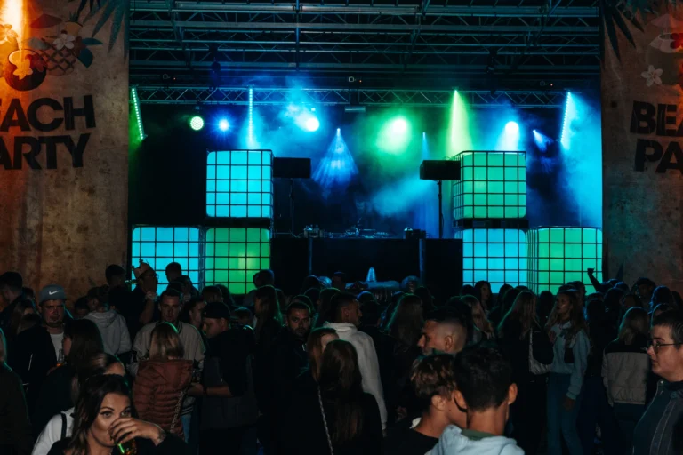 Gäste stehen vor der Bühne zwischen Beachparty-Bannern; grüne und blaue Scheinwerfer beleuchten quadratische Lichtflächen.